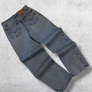 May include: Light blue denim jeans with classic five-pocket styling. The jeans feature a faded wash and a leather patch on the back waistband. The back pockets have a signature curved stitch design.