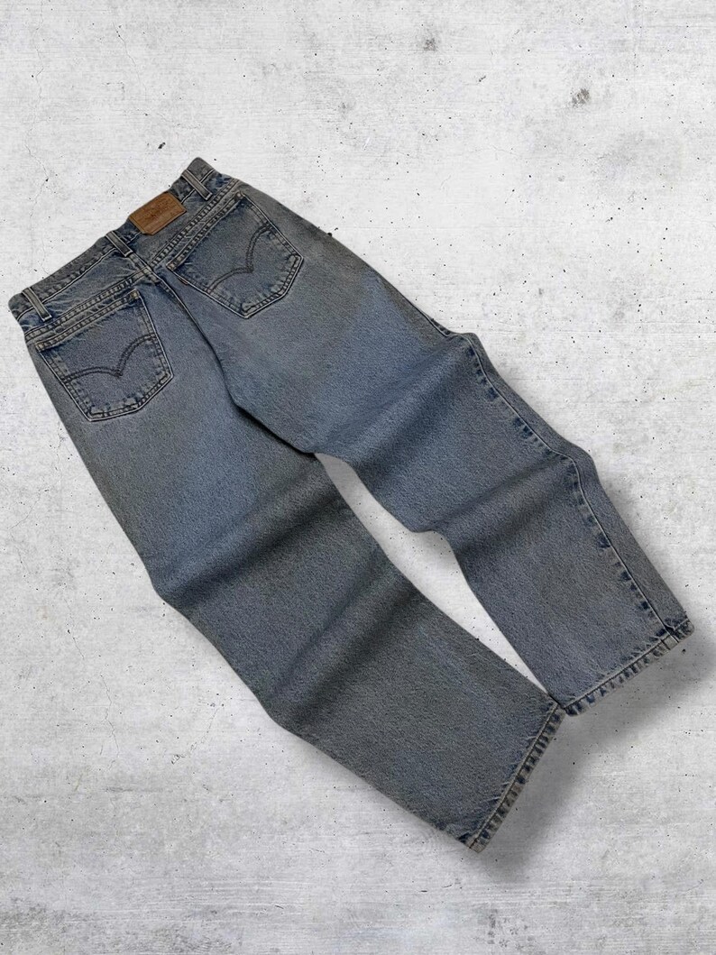 May include: Light blue denim jeans with classic five-pocket styling. The jeans feature a faded wash and a leather patch on the back waistband. The back pockets have a signature curved stitch design.