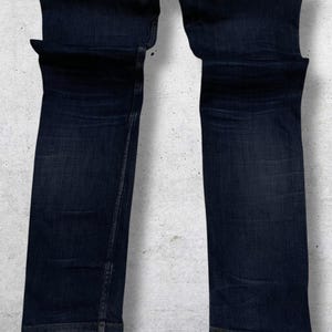 Op de afbeelding: Donkerblauwe denim jeans. De jeans zijn opgevouwen en gepresenteerd tegen een lichtgrijze achtergrond. De jeans hebben een klassiek ontwerp met vijf zakken en een rechte pijp. De denim heeft subtiele vervagingen en whiskers.