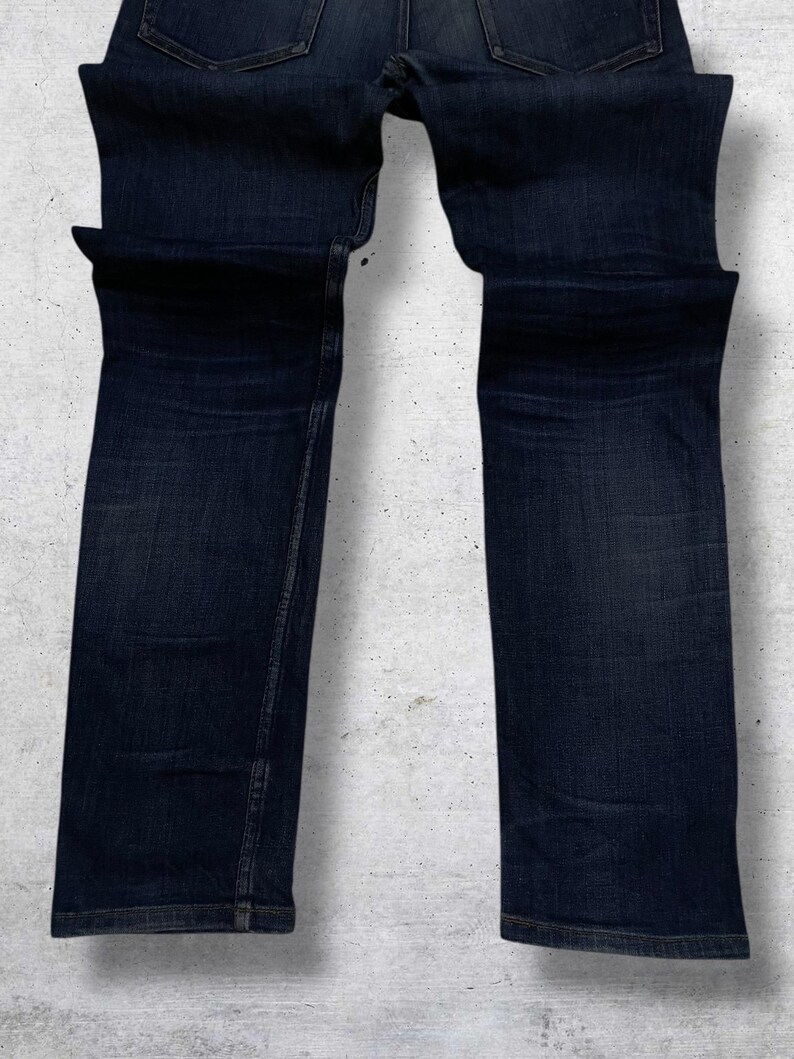 Op de afbeelding: Donkerblauwe denim jeans. De jeans zijn opgevouwen en gepresenteerd tegen een lichtgrijze achtergrond. De jeans hebben een klassiek ontwerp met vijf zakken en een rechte pijp. De denim heeft subtiele vervagingen en whiskers.
