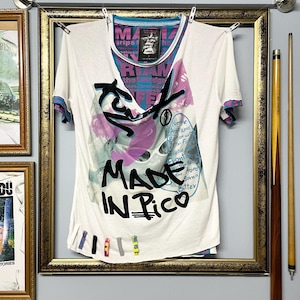 Könnte beinhalten: Weißes T-Shirt mit einem grafischen Design mit dem Text "MADE IN PICO" in Schwarz. Das Shirt hat einen V-Ausschnitt und kurze Ärmel mit blauen und lila Akzenten. Das Shirt hängt an einer Wäscheleine in einem goldgerahmten Bild.