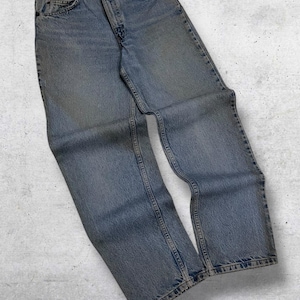 May include: Light blue denim jeans with a classic five-pocket design. The jeans have a button and zipper closure and show signs of wear. The denim is faded, giving the jeans a vintage look. The jeans are laid flat on a light gray surface.