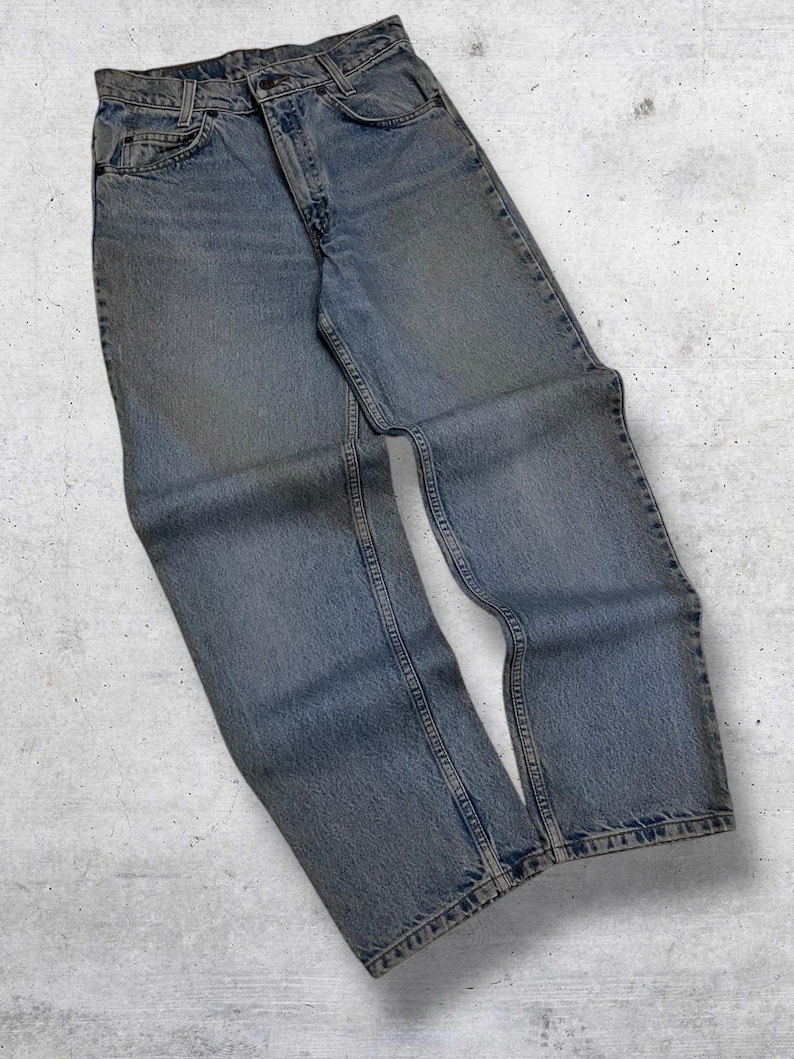 May include: Light blue denim jeans with a classic five-pocket design. The jeans have a button and zipper closure and show signs of wear. The denim is faded, giving the jeans a vintage look. The jeans are laid flat on a light gray surface.