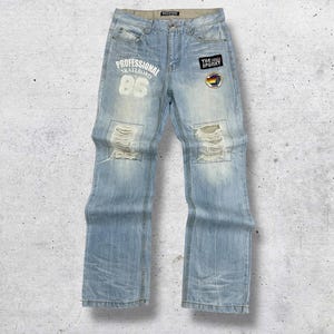 May include: Light blue denim jeans with distressed details. Features the text "PROFESSIONAL SKATEBOARD 86" on the left leg and patches on the right leg. The jeans have a relaxed fit and a faded wash.