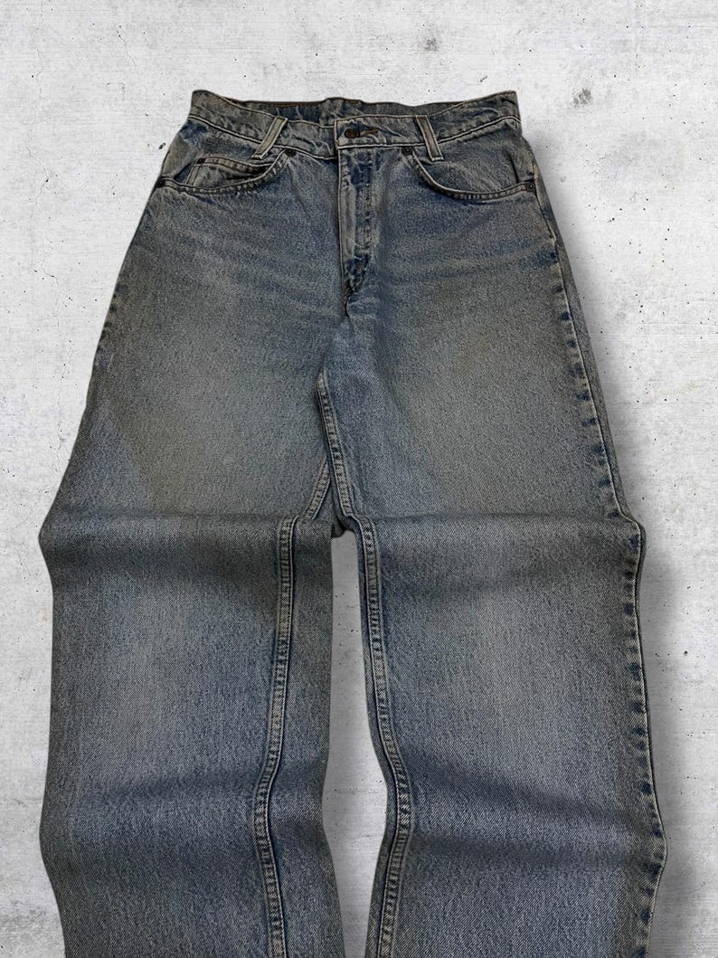 May include: Light blue denim jeans with a classic five-pocket design. The jeans have a faded wash and a straight leg cut. The image shows the jeans laid flat, showcasing the front pockets, button closure, and belt loops. The denim appears worn.