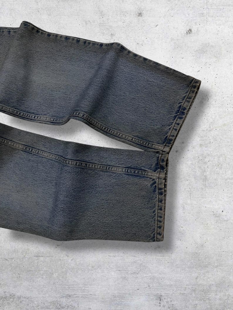 May include: Close-up of a pair of faded blue jeans. The denim pants are folded, showcasing the texture and stitching details. The jeans have a classic five-pocket design and a slightly worn appearance.
