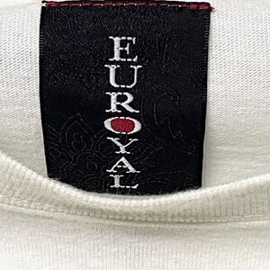 May include: Close-up of a white t-shirt with a black label. The label features the word "EUROYAL" in white capital letters, with a red circle in the middle of the word. The label has a decorative floral pattern.
