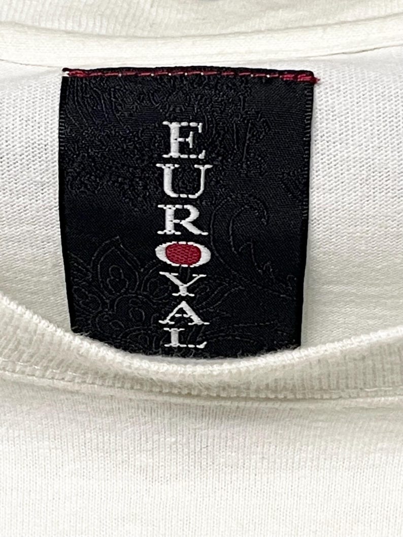 May include: Close-up of a white t-shirt with a black label. The label features the word "EUROYAL" in white capital letters, with a red circle in the middle of the word. The label has a decorative floral pattern.