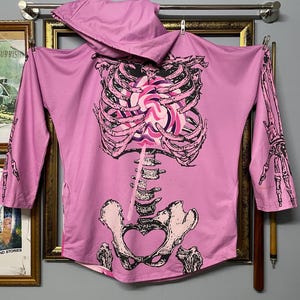 Puede incluir: Una sudadera con capucha rosa claro con un gráfico de esqueleto. El diseño del esqueleto incluye una caja torácica, columna vertebral y pelvis, con un corazón de caramelo colorido en el pecho. Las mangas presentan gráficos de manos de esqueleto.