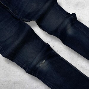 Op de afbeelding: Donkerblauwe denim jeans met een beschadigd kniedetail. De jeans is opgevouwen, wat de textuur en kleurvariaties van de stof laat zien. De algehele uitstraling is casual en gedragen.