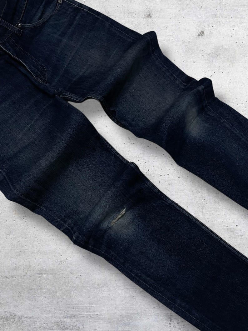 Op de afbeelding: Donkerblauwe denim jeans met een beschadigd kniedetail. De jeans is opgevouwen, wat de textuur en kleurvariaties van de stof laat zien. De algehele uitstraling is casual en gedragen.