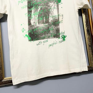 May include: White t-shirt with a green and grey graphic print. The print features a grayscale image of a cave or tunnel with green text overlay. The text includes phrases like "It's up to human beings to protect" and "with good people's hands."