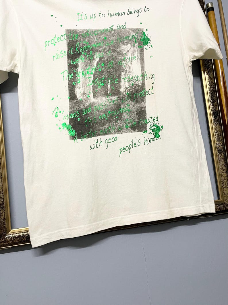 May include: White t-shirt with a green and grey graphic print. The print features a grayscale image of a cave or tunnel with green text overlay. The text includes phrases like "It's up to human beings to protect" and "with good people's hands."