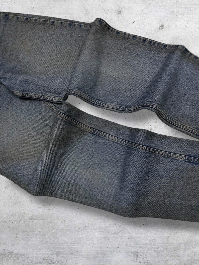 May include: Faded blue denim jeans, folded and laid flat. The jeans have a classic five-pocket design with visible stitching and a worn appearance. The fabric has a slightly textured look, suggesting a vintage or well-loved item.