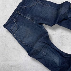 以下が含まれることがあります： クラシックな5ポケットデザインの濃い青色のデニムジーンズ。ジーンズはボタンとジッパーで開閉し、ベルトループがあり、使い古したようなウォッシュ加工が施されています。ジーンズは明るい灰色の表面に平らに置かれています。