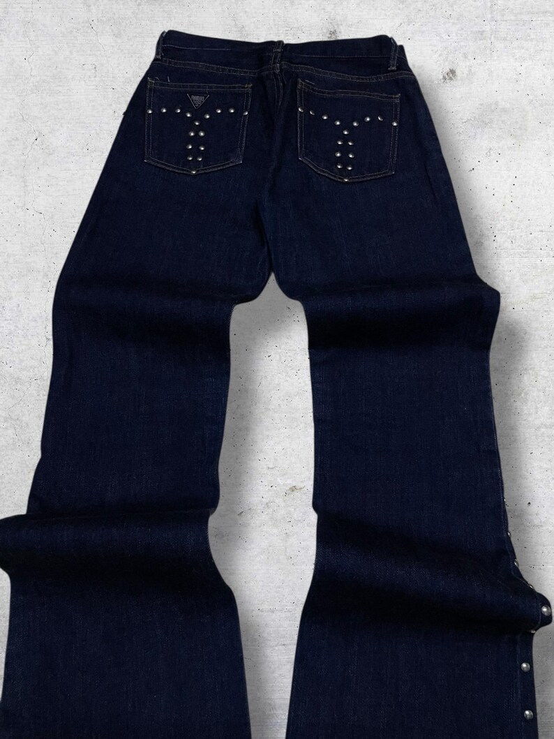 Puede incluir: Vaqueros de mezclilla azul oscuro con pernera acampanada. Los bolsillos traseros presentan un dise&ntilde;o decorativo con tachuelas plateadas. Los vaqueros tienen un dise&ntilde;o cl&aacute;sico de cinco bolsillos y cierre de bot&oacute;n y cremallera.
