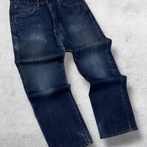 May include: Dark wash denim jeans with a classic five-pocket design. The jeans have a straight leg cut and a button and zipper closure. The denim has a faded appearance, with subtle wear marks. The jeans are laid flat on a textured surface.