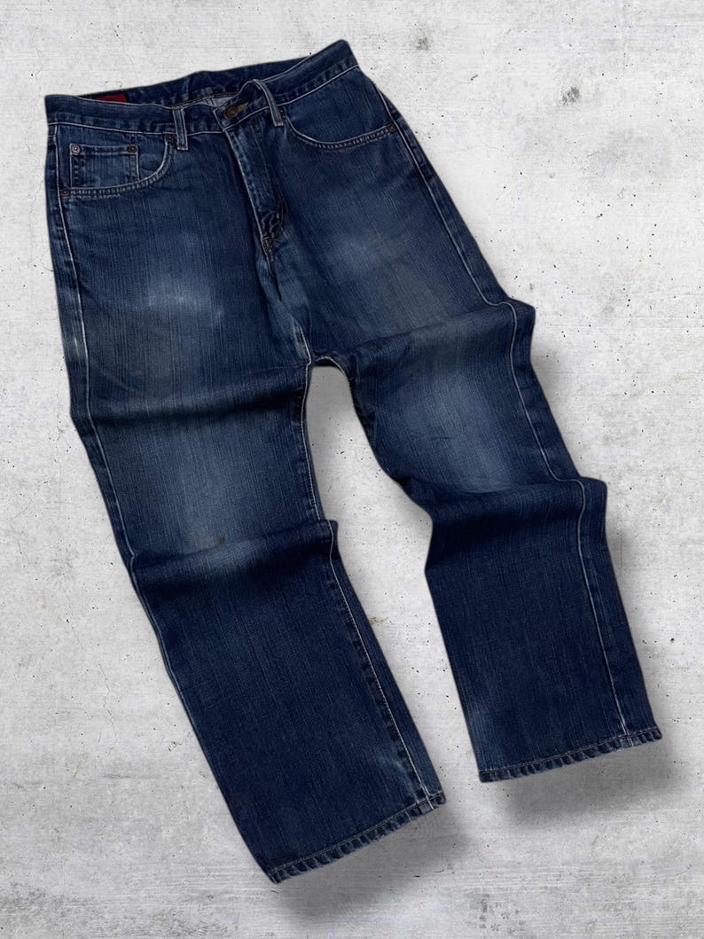 May include: Dark wash denim jeans with a classic five-pocket design. The jeans have a straight leg cut and a button and zipper closure. The denim has a faded appearance, with subtle wear marks. The jeans are laid flat on a textured surface.
