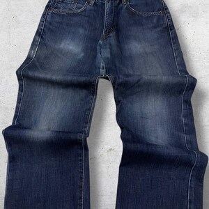 May include: Dark wash denim jeans with a classic five-pocket design. The jeans have a straight leg cut and a button and zipper closure. The denim has subtle fading and wear marks, giving them a vintage look. The jeans are laid flat on a light gray surface.