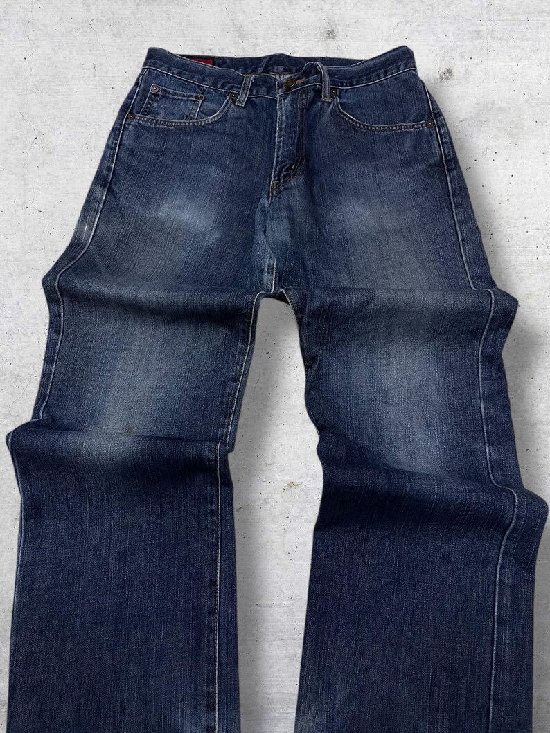May include: Dark wash denim jeans with a classic five-pocket design. The jeans have a straight leg cut and a button and zipper closure. The denim has subtle fading and wear marks, giving them a vintage look. The jeans are laid flat on a light gray surface.