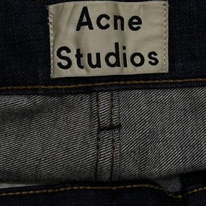 Op de afbeelding: Close-up van een donkerblauwe denim kledingstuk met het label "Acne Studios". Het rechthoekige label heeft zwarte tekst op een lichte achtergrond. Zichtbare stiksels en stoftextuur.