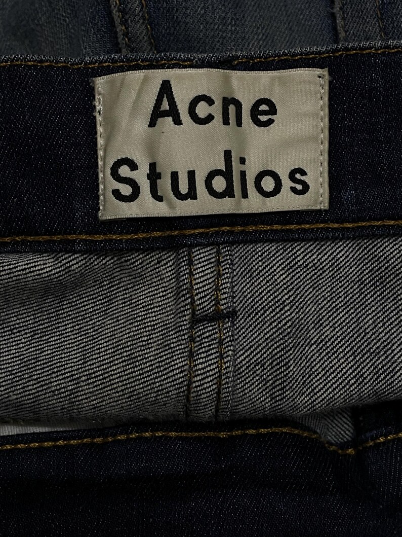 Op de afbeelding: Close-up van een donkerblauwe denim kledingstuk met het label "Acne Studios". Het rechthoekige label heeft zwarte tekst op een lichte achtergrond. Zichtbare stiksels en stoftextuur.