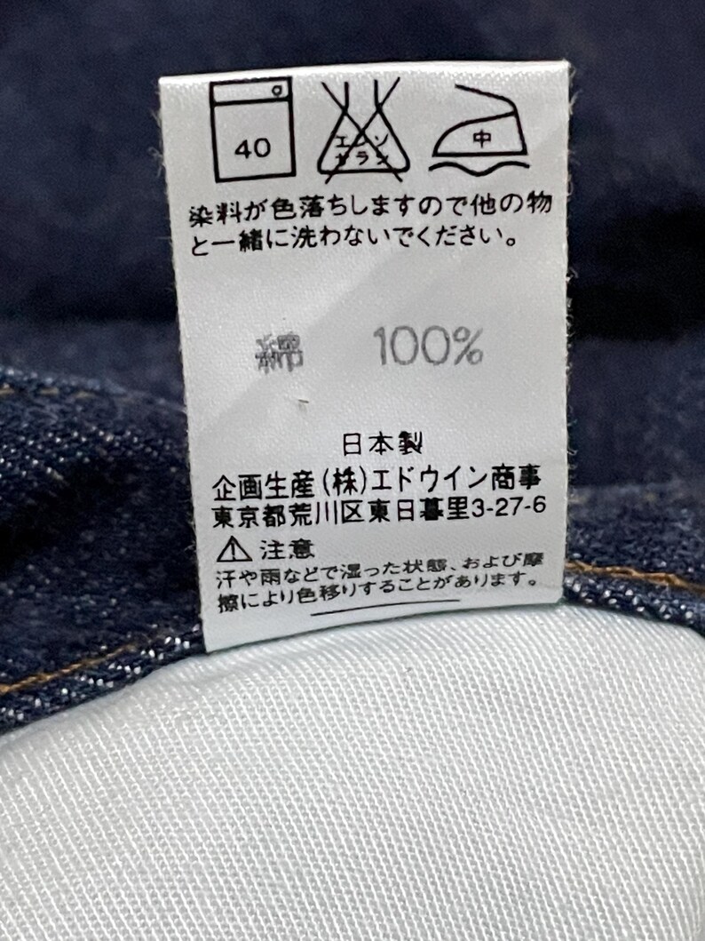Puede incluir: Primer plano de una etiqueta blanca con texto y s&iacute;mbolos japoneses, probablemente una etiqueta de cuidado de la ropa. La etiqueta incluye instrucciones de lavado, contenido de la tela (100% algod&oacute;n) e informaci&oacute;n de fabricaci&oacute;n.