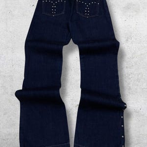 Puede incluir: Vaqueros de mezclilla azul oscuro con pernera acampanada. Los bolsillos traseros est&aacute;n adornados con tachuelas plateadas. Los vaqueros tienen cierre de bot&oacute;n y cremallera y tachuelas plateadas a lo largo de las costuras laterales. Los vaqueros se muestran sobre un fondo gris claro y blanco.