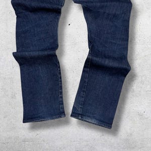 以下が含まれることがあります： 濃い青色のデニムジーンズ。ジーンズは、クラシックな5ポケットのデザインとストレートレッグカットです。デニムはわずかに色あせた外観で、微妙な摩耗の跡があります。ジーンズは裾が折り返されています。背景は、明るいグレーのテクスチャード加工された表面です。