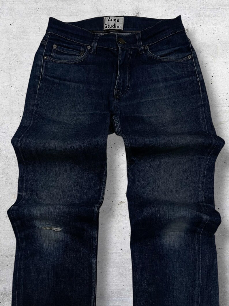 Op de afbeelding: Donkerblauwe denim jeans met een beschadigde knie. De jeans hebben een klassiek ontwerp met vijf zakken en een knoop- en ritssluiting. De merknaam "Acne Studios" is zichtbaar op de tailleband.