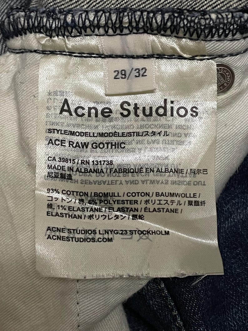 Op de afbeelding: Close-up van een wit label van Acne Studios jeans. Het label toont maat 29/32, de merknaam en de stijl "ACE RAW GOTHIC". Het geeft ook de materialen weer: 93% katoen, 6% polyester en 1% elastaan.