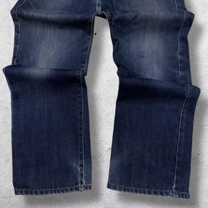May include: Dark blue denim jeans with a classic five-pocket design. The jeans have a straight leg cut and show subtle fading and wear. The stitching is visible in a lighter thread color, and the fabric has a textured appearance.