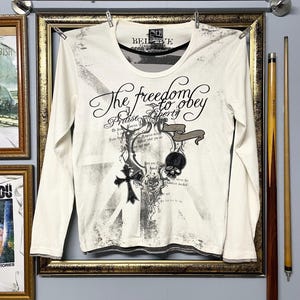 May include: Cream-colored long-sleeve shirt with a crew neck and graphic design. The design includes a cross, skull, and the text "The freedom to obey". The shirt is hanging on a clothesline.