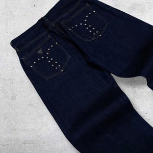 Puede incluir: Vaqueros de mezclilla azul oscuro con tachuelas plateadas en forma de Y en los bolsillos traseros. Los vaqueros presentan un dise&ntilde;o cl&aacute;sico de cinco bolsillos y un peque&ntilde;o parche con el logotipo en el bolsillo trasero.