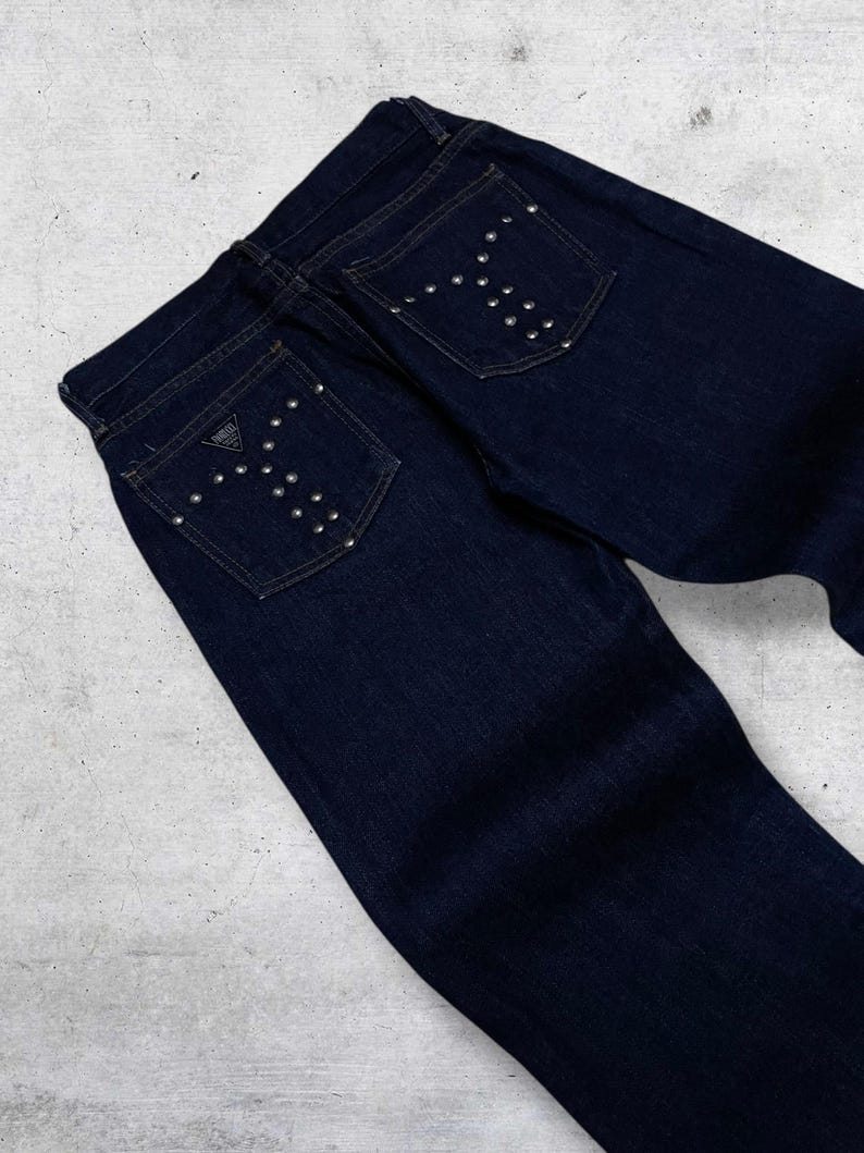 Puede incluir: Vaqueros de mezclilla azul oscuro con tachuelas plateadas en forma de Y en los bolsillos traseros. Los vaqueros presentan un dise&ntilde;o cl&aacute;sico de cinco bolsillos y un peque&ntilde;o parche con el logotipo en el bolsillo trasero.