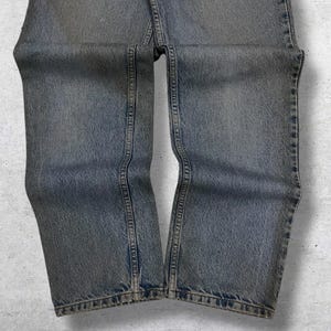 May include: Faded blue denim jeans with a straight leg cut. The jeans show signs of wear and have visible seams and stitching details. The denim has a slightly distressed appearance, with subtle fading and a vintage aesthetic.