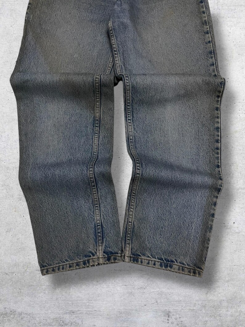 May include: Faded blue denim jeans with a straight leg cut. The jeans show signs of wear and have visible seams and stitching details. The denim has a slightly distressed appearance, with subtle fading and a vintage aesthetic.