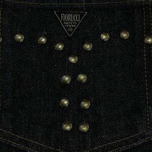 Puede incluir: Primer plano de un bolsillo de mezclilla azul oscuro con tachuelas met&aacute;licas doradas dispuestas en un patr&oacute;n decorativo. El bolsillo presenta el logotipo "FIORUCCI SAFETY JEANS" en blanco. La costura es dorada.