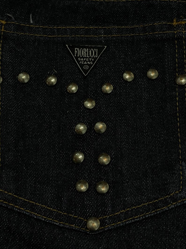 Puede incluir: Primer plano de un bolsillo de mezclilla azul oscuro con tachuelas met&aacute;licas doradas dispuestas en un patr&oacute;n decorativo. El bolsillo presenta el logotipo "FIORUCCI SAFETY JEANS" en blanco. La costura es dorada.