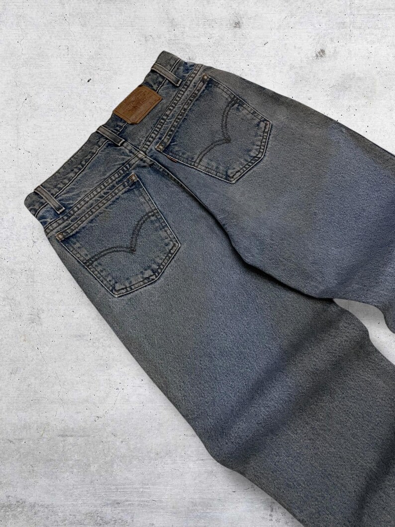 May include: Faded blue denim jeans with classic five-pocket styling. Features a brown leather patch and the iconic Levi's back pocket stitching. The jeans are pre-worn, showing signs of wear.