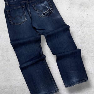 May include: Dark blue denim jeans with a distressed look, featuring a worn patch on the back pocket. The jeans have a classic five-pocket design and a leather patch with the brand name "EDWIN".