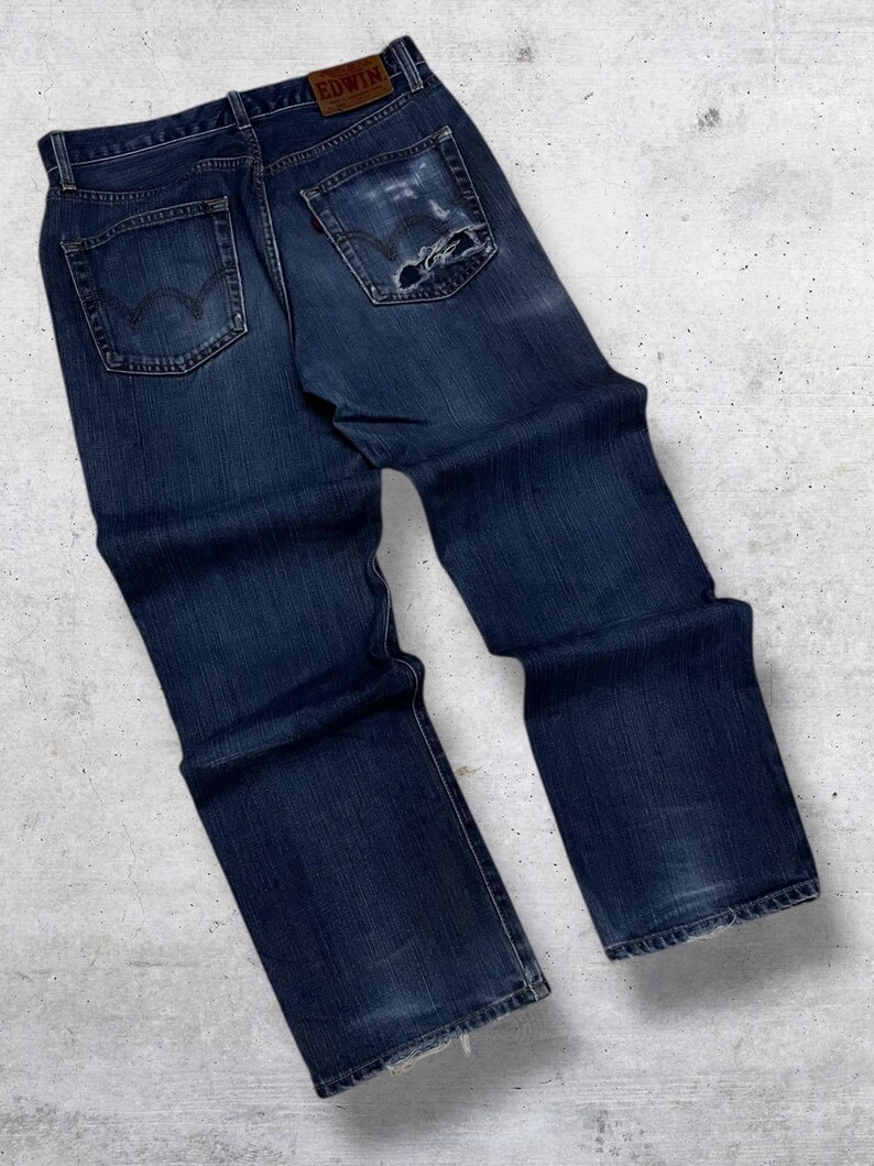 May include: Dark blue denim jeans with a distressed look, featuring a worn patch on the back pocket. The jeans have a classic five-pocket design and a leather patch with the brand name "EDWIN".