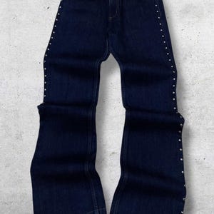 Puede incluir: Vaqueros de mezclilla azul oscuro con detalles de tachuelas plateadas a lo largo de las costuras laterales y los bolsillos delanteros. Los vaqueros tienen una abertura acampanada en la pierna y un dise&ntilde;o cl&aacute;sico de cinco bolsillos. Los vaqueros se muestran sobre un fondo gris claro.