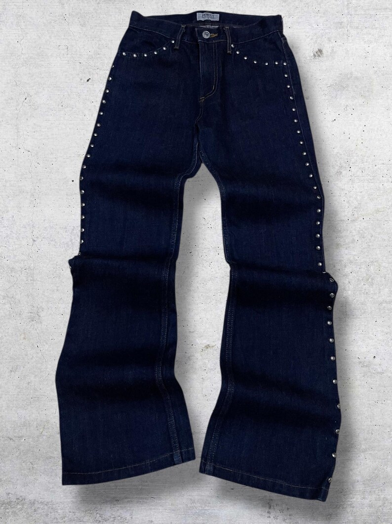 Puede incluir: Vaqueros de mezclilla azul oscuro con detalles de tachuelas plateadas a lo largo de las costuras laterales y los bolsillos delanteros. Los vaqueros tienen una abertura acampanada en la pierna y un dise&ntilde;o cl&aacute;sico de cinco bolsillos. Los vaqueros se muestran sobre un fondo gris claro.