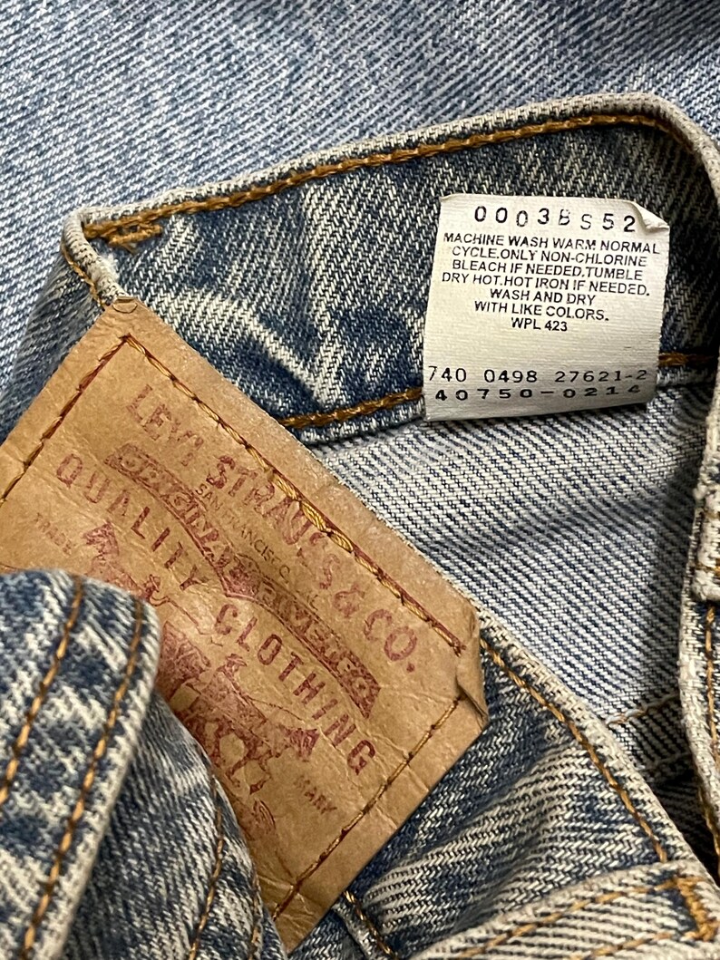 May include: Close-up of a denim garment with a brown leather tag and a white tag. The brown tag reads "LEVI STRAUSS & CO. QUALITY CLOTHING". The white tag has washing instructions. The denim is blue with visible stitching.