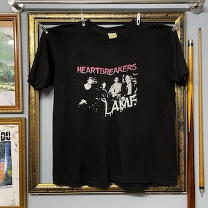Puede incluir: Camiseta negra con la inscripción rosa "HEARTBREAKERS" sobre una imagen de banda y la palabra blanca "LAME". La camiseta está colgada en un tendedero dentro de un marco dorado.