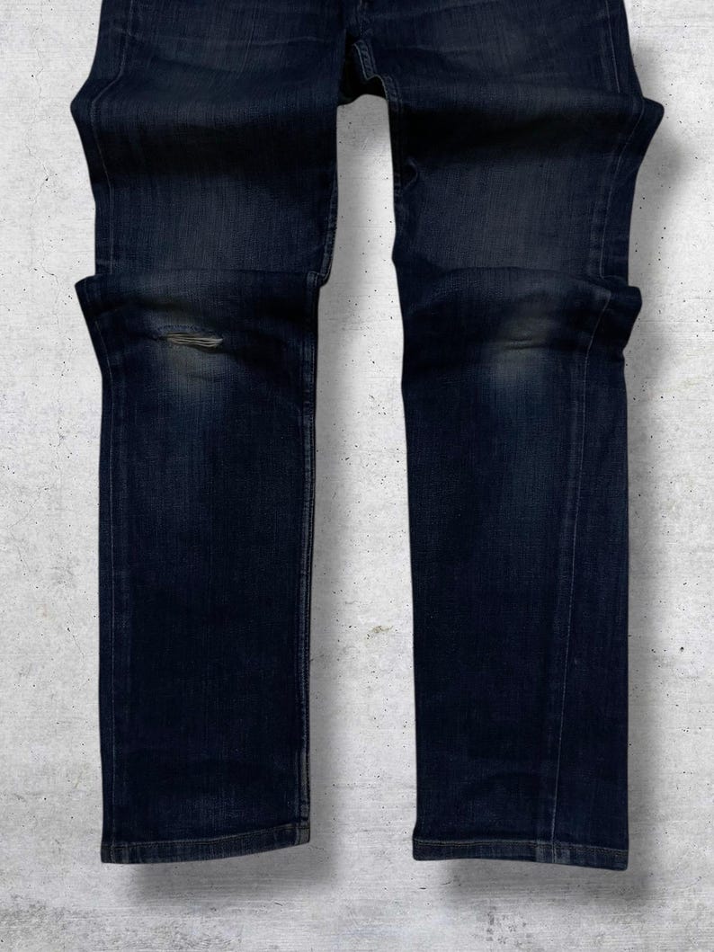 Op de afbeelding: Donkerblauwe denim jeans met een beschadigd kniedetail. De jeans hebben een klassiek vijf-zakken ontwerp en een rechte pijp. De denim heeft een vervaagde uitstraling, met subtiele kleurvariaties.