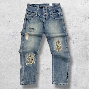 May include: Light blue denim jeans with distressed details, including a knee-level tear and decorative patches. The patches feature floral patterns and are outlined with white stitching. The jeans have a classic five-pocket design.