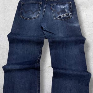 May include: Dark blue denim jeans with a distressed pocket. The jeans have a classic five-pocket design, a red tag, and the brand name "EDWIN" on the back. The jeans are slightly faded and show signs of wear.