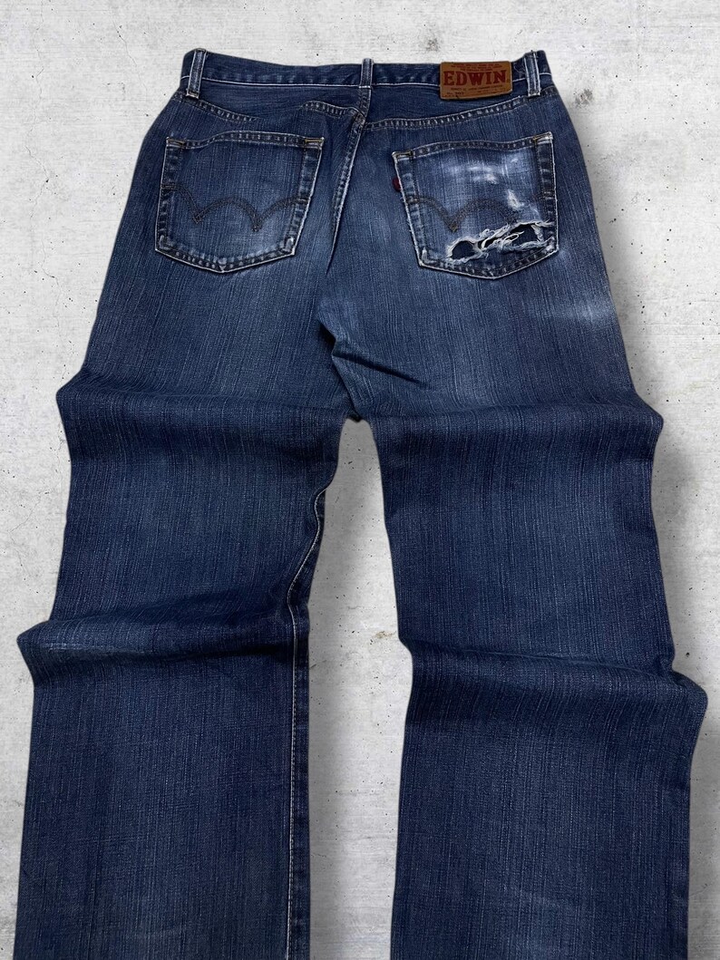 May include: Dark blue denim jeans with a distressed pocket. The jeans have a classic five-pocket design, a red tag, and the brand name "EDWIN" on the back. The jeans are slightly faded and show signs of wear.
